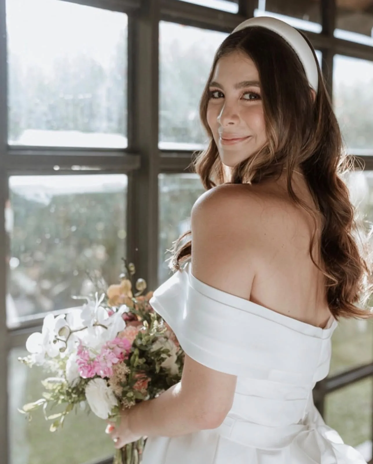Novia mirando por encima del hombro con ramo de flores junto a una ventana