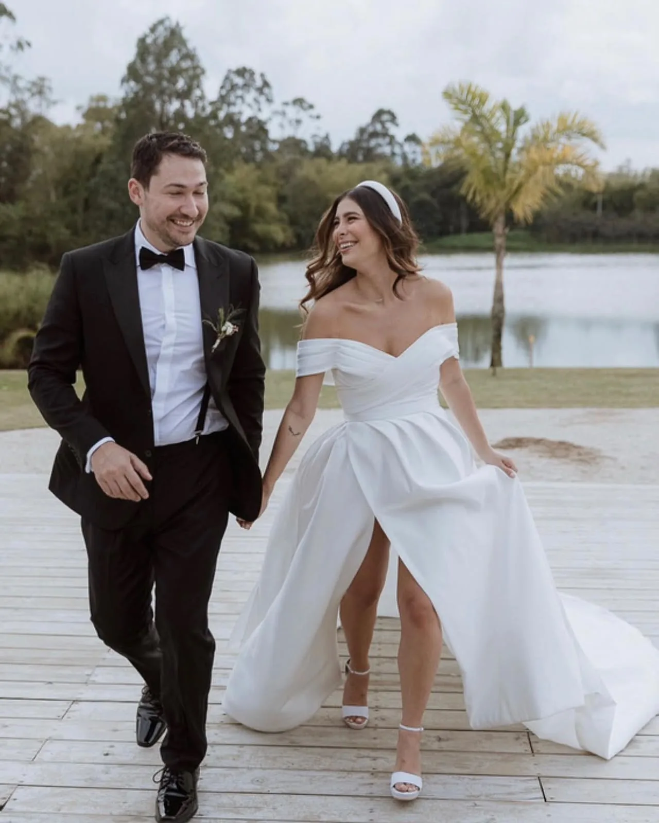 Pareja de novios caminando junto a un lago al atardecer, novia con vestido escotado y falda con abertura