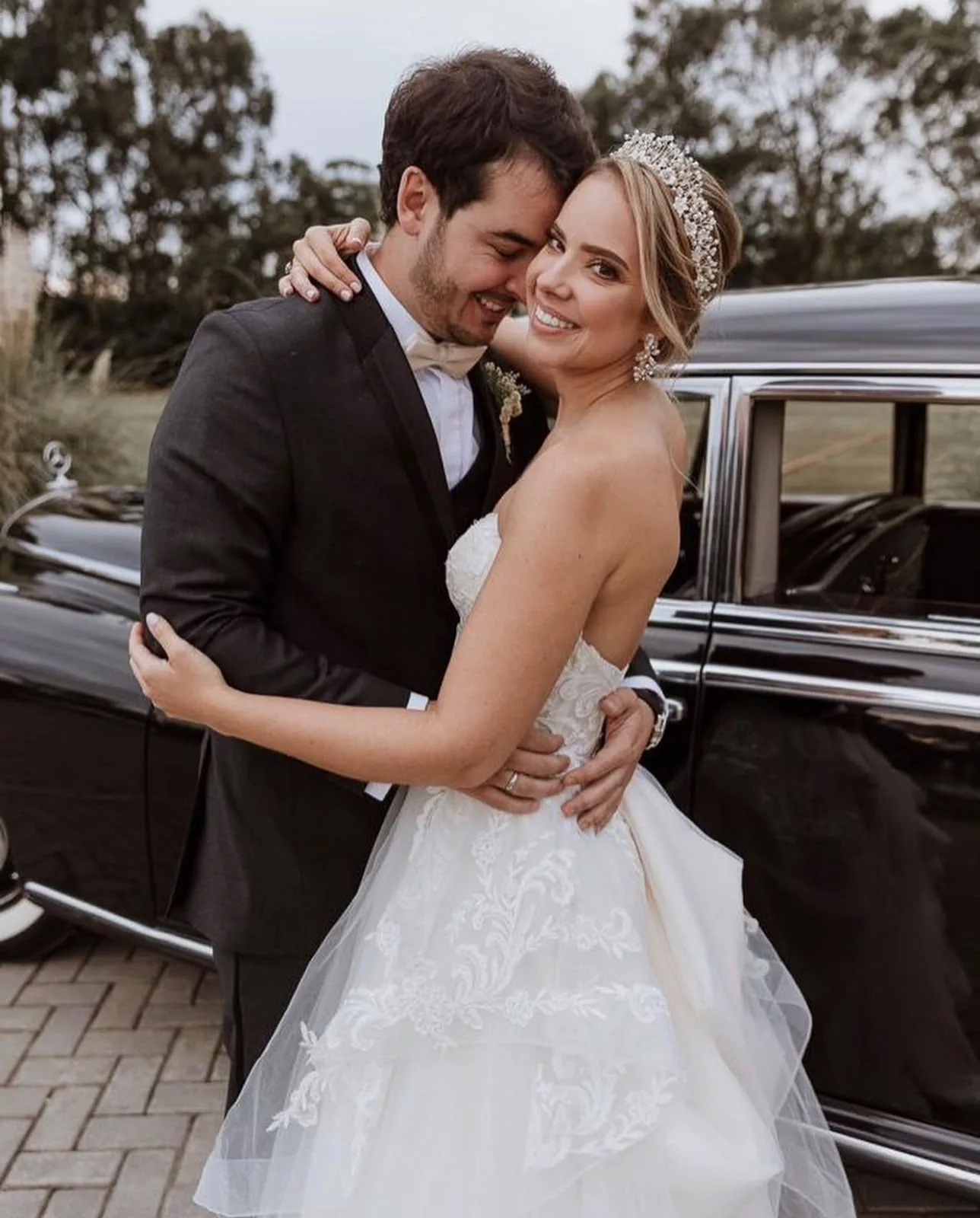 Pareja de novios abrazados junto a un auto clásico, novia con tiara de cristales y vestido de encaje